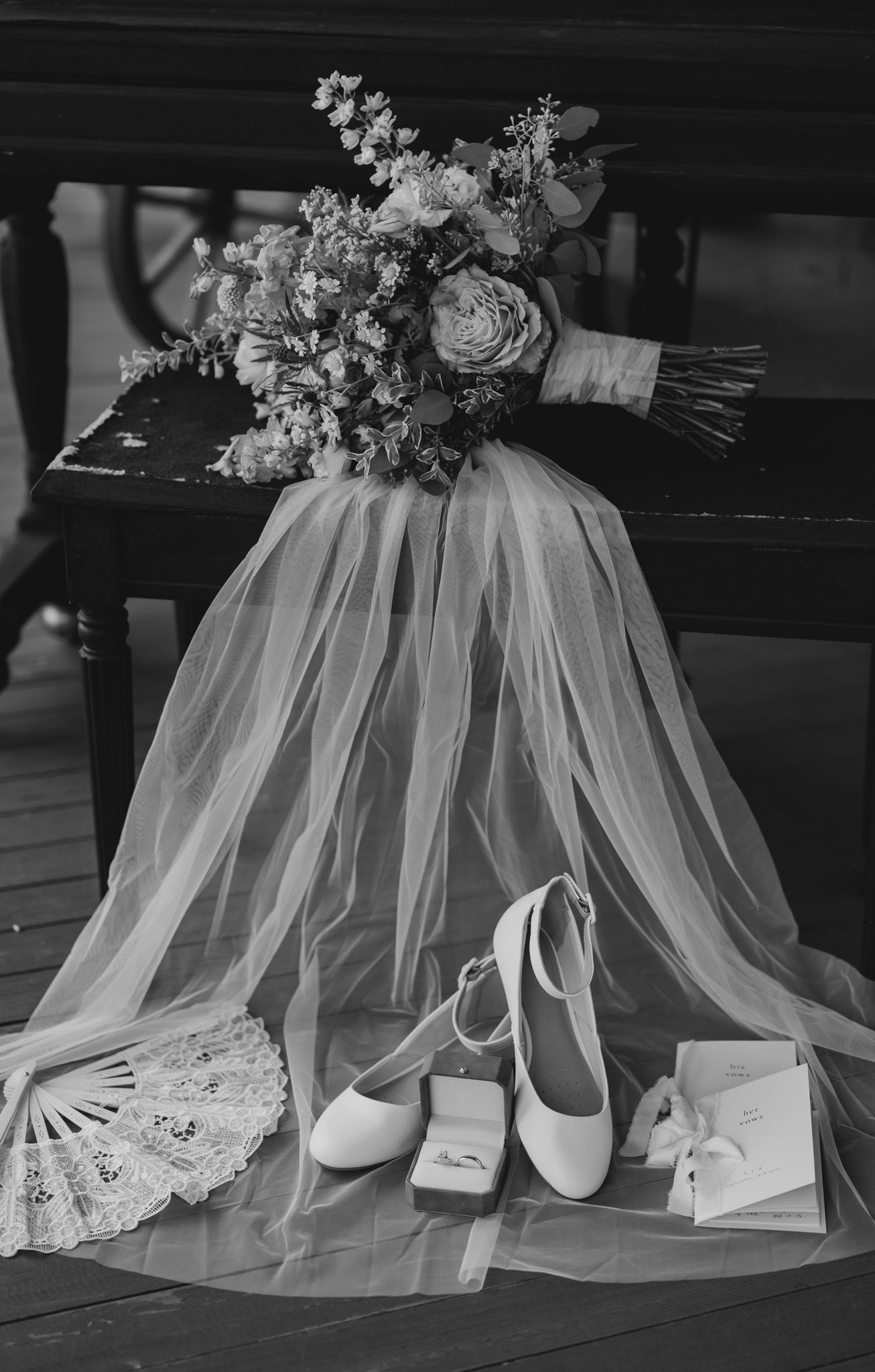 Wedding details featuring a bridal bouquet, veil, white shoes, and accessories arranged on a dark bench.