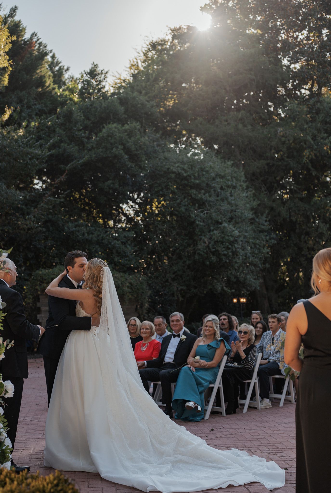 A romantic outdoor wedding ceremony with guests seated on white chairs as sunlight filters through trees in the background.
