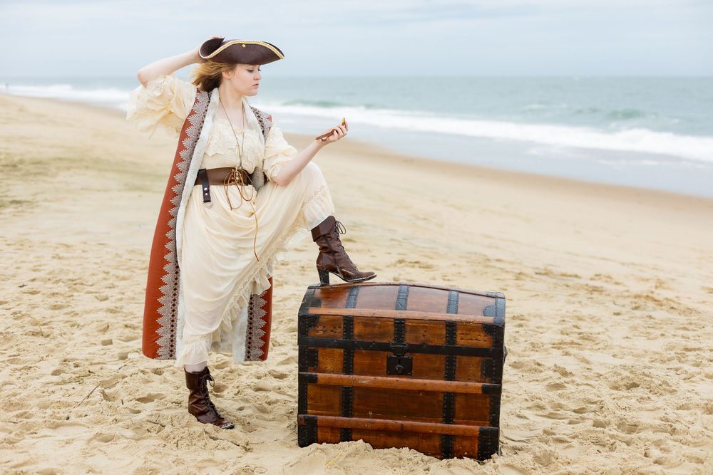 A pirate puts one leg up on a treasure chest while looking at a compass
