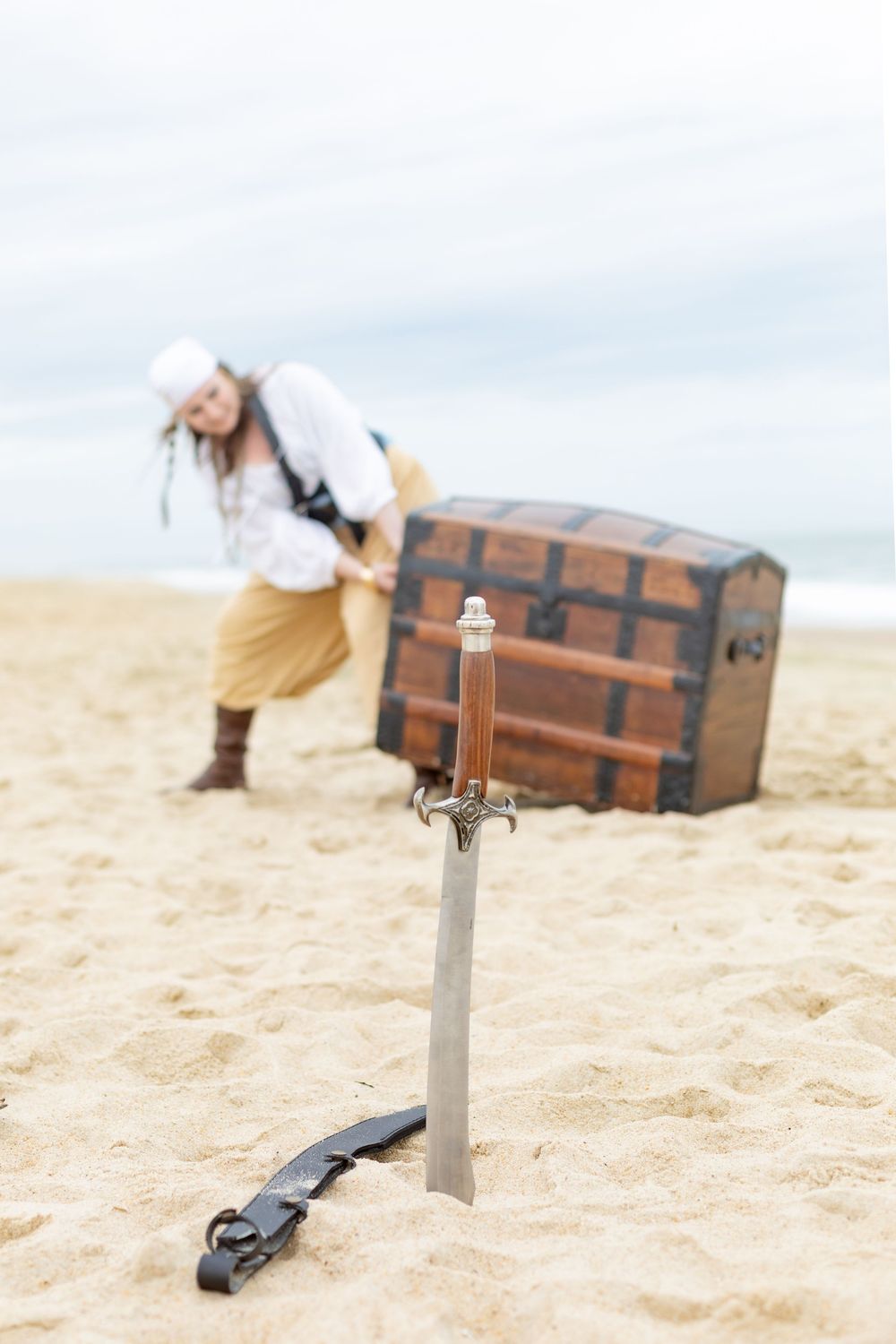 Pirate lugging a big chest in the background while a swont sticks out in the sand int he foreground