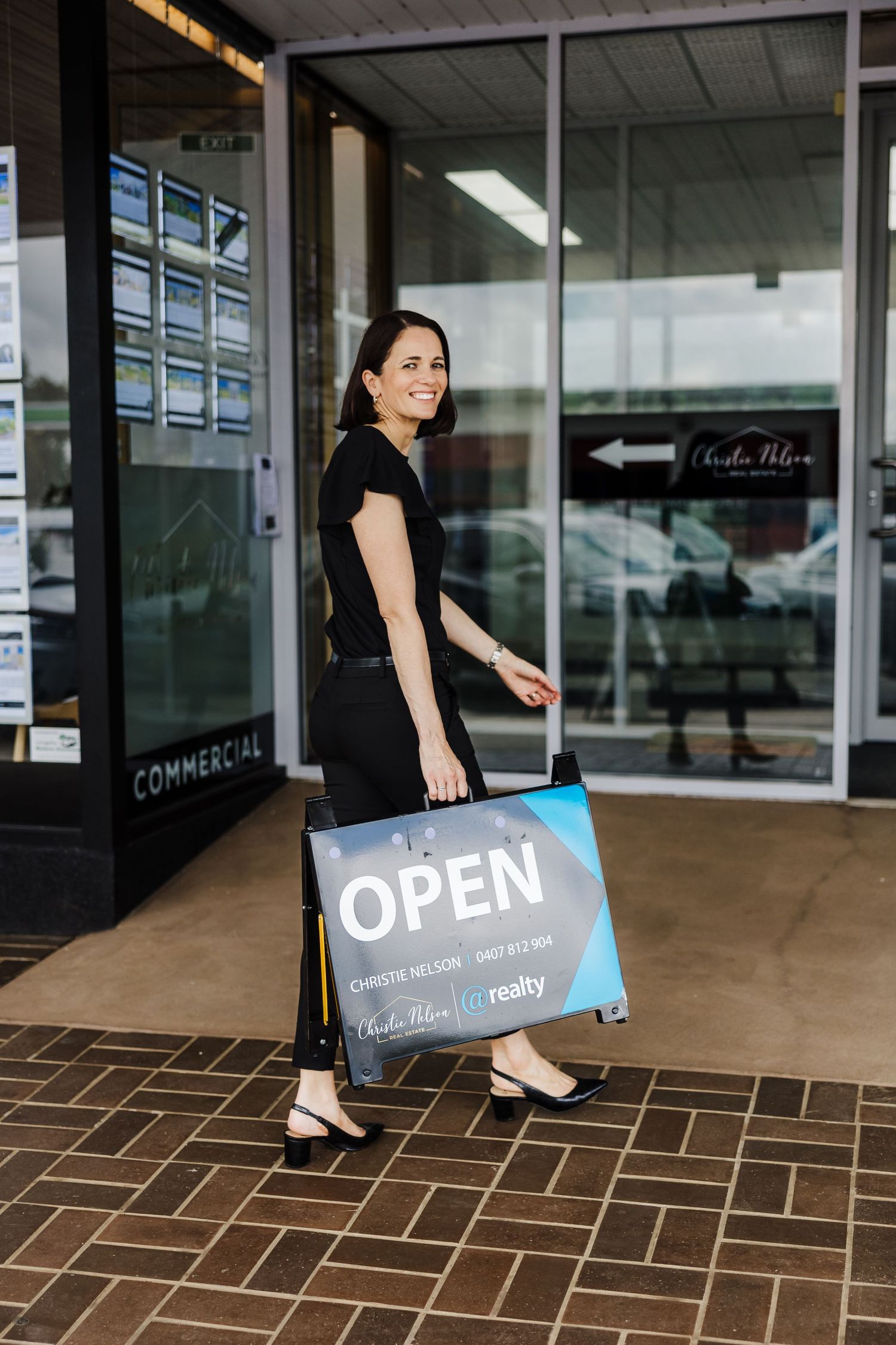 Business owner in black attire places an open sign outside a commercial storefront with brick pavers and glass windows.t. Business Headshots