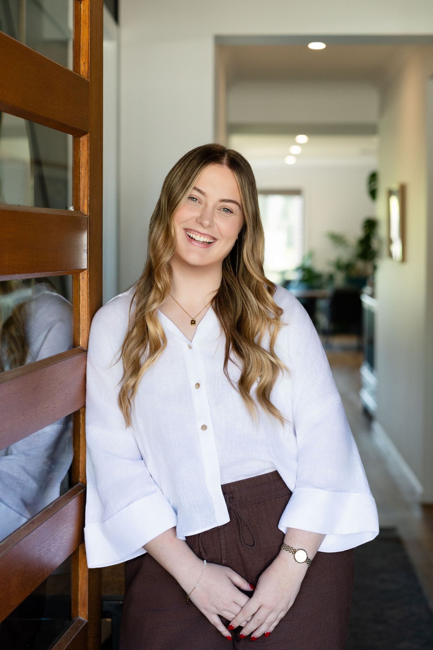 Professional portrait in a bright hallway featuring a white button-down blouse and brown skirt with warm interior lighting.