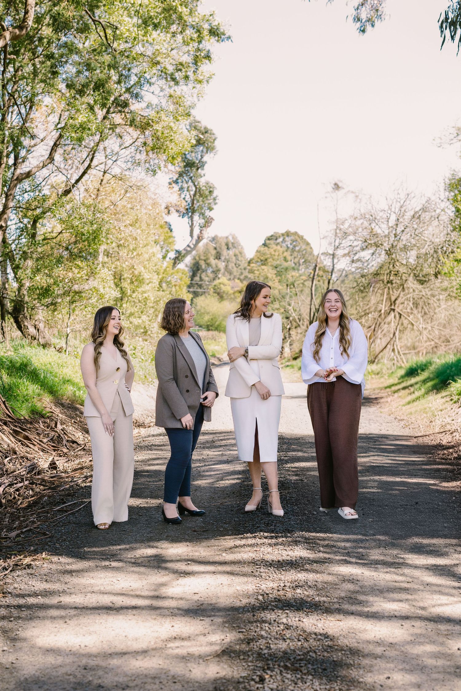 A group of people stand together on a sunlit dirt path surrounded by trees and natural scenery.