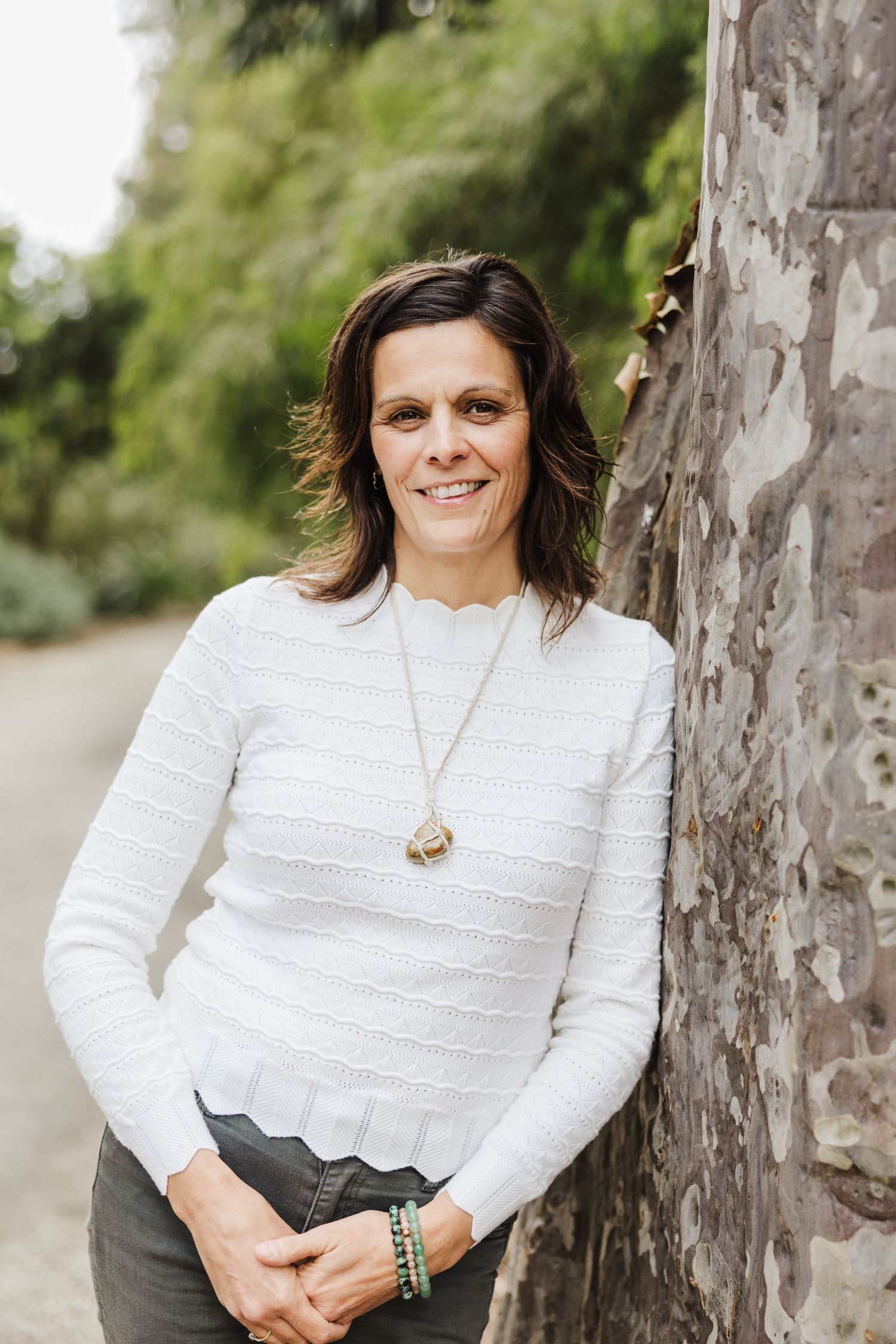 Portrait of someone in a white sweater leaning against a tree trunk outdoors.