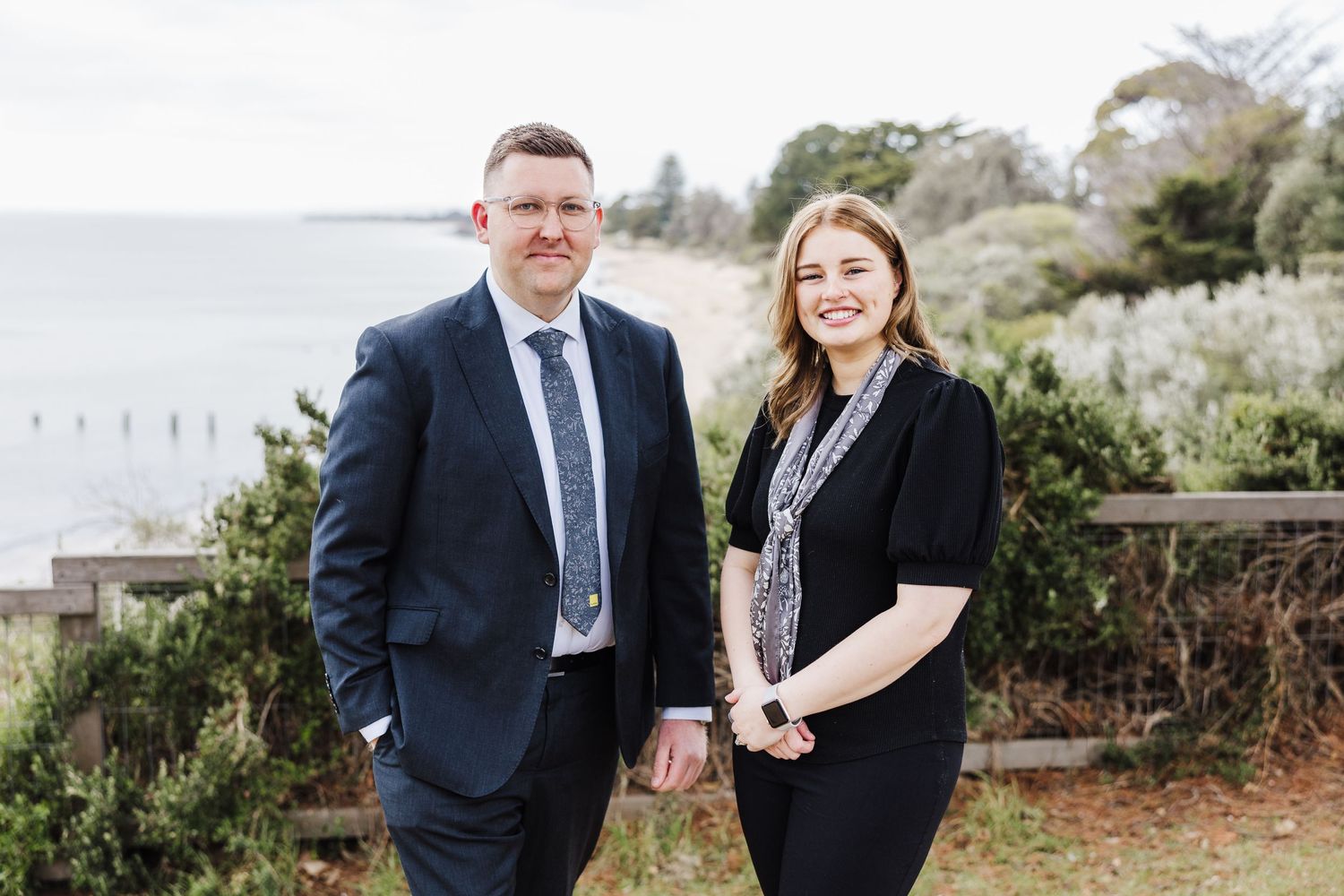 Two professionals in business attire pose together on a coastal overlook.