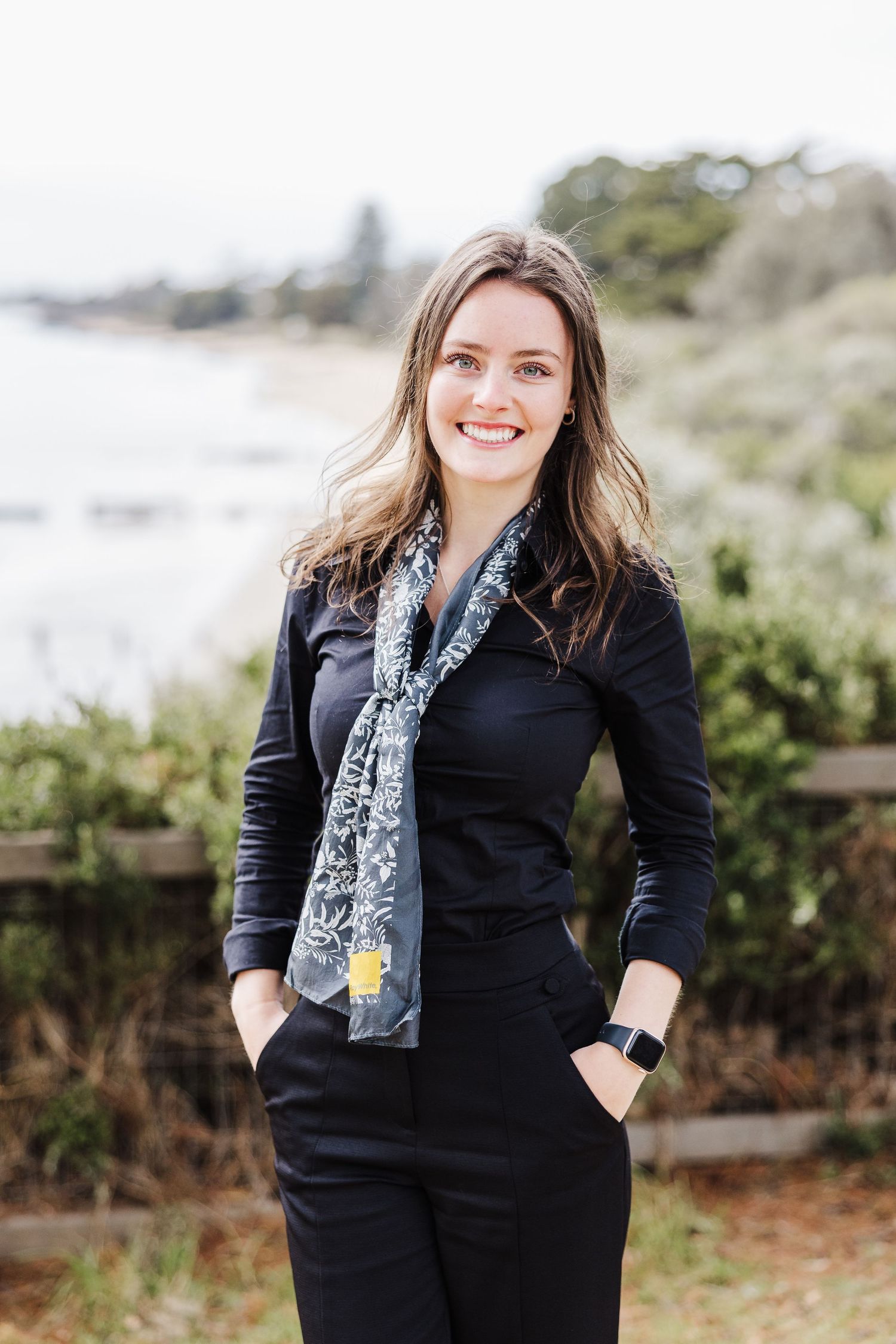 A person in a black outfit with patterned scarf stands outdoors against a coastal backdrop.