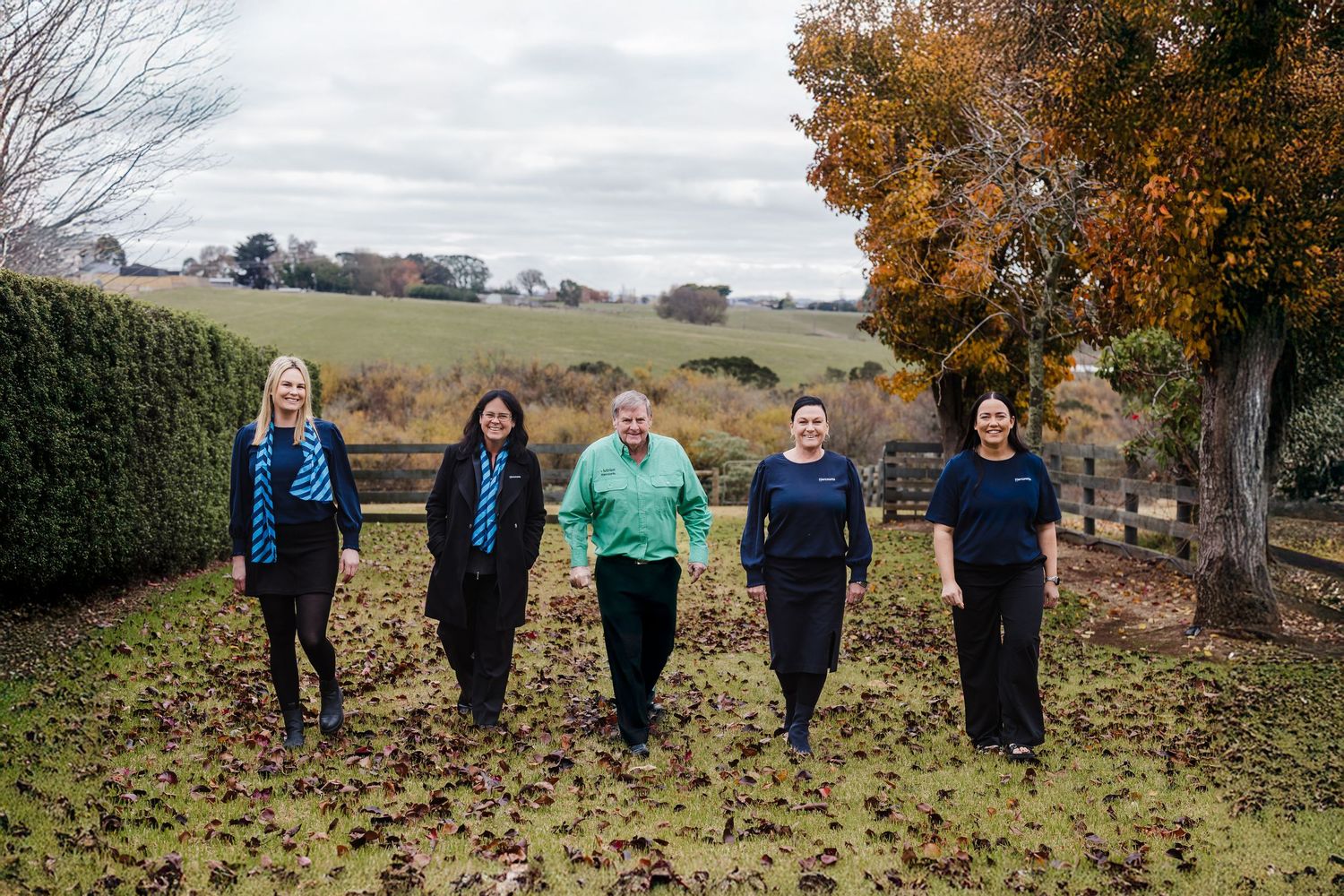 A group of people stand in a line against an autumn landscape with bare trees and rolling hills in the background.