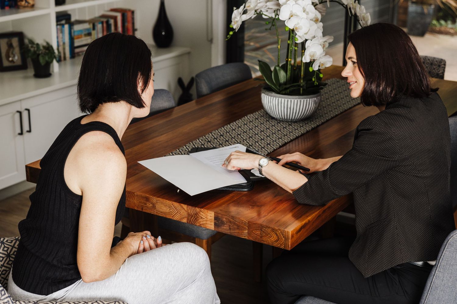 Two people sit at a wooden table having a business meeting with papers and an orchid centerpiece.