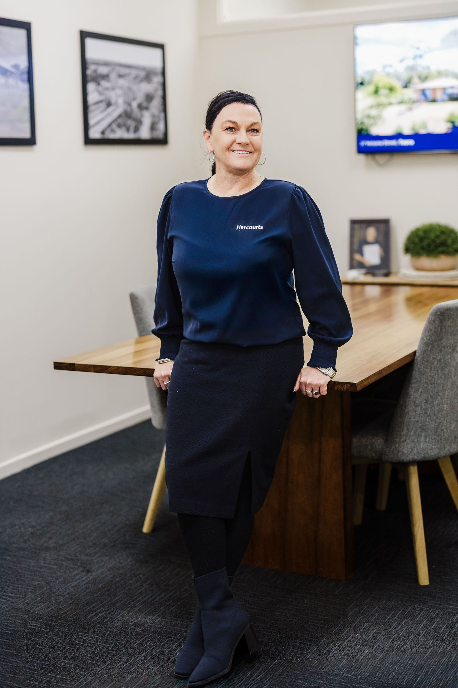 Person in navy dress stands in modern office space with wooden tables and gray chairs against white walls.