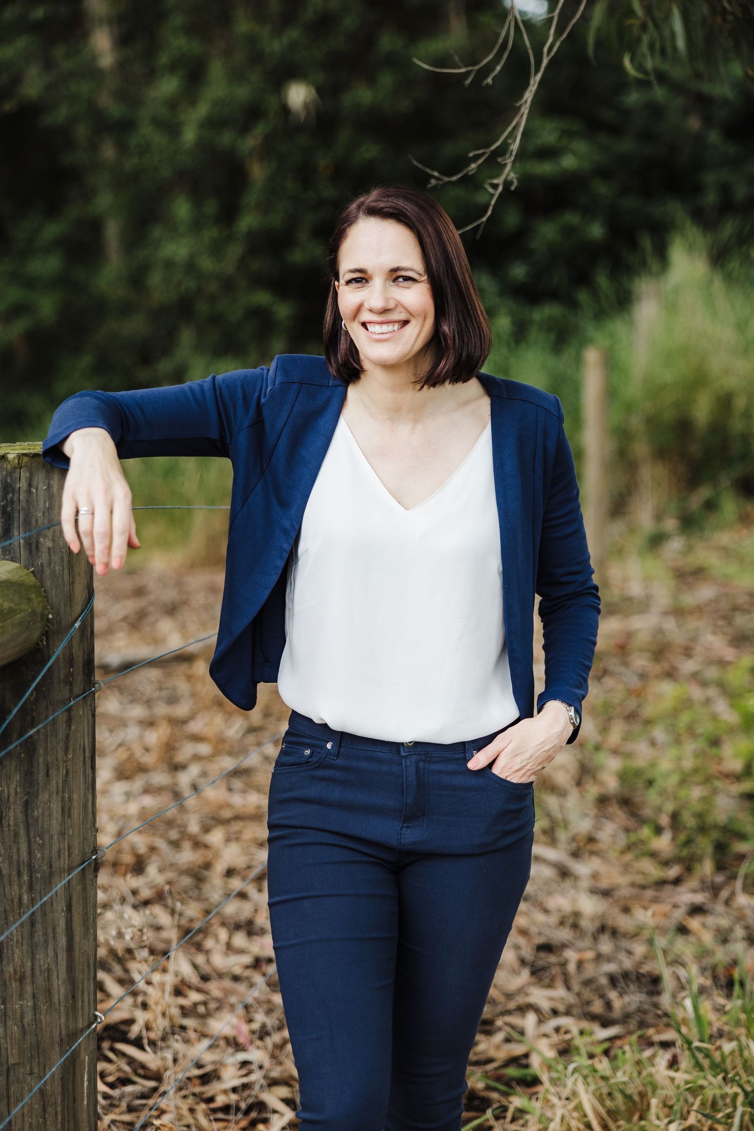 Relaxed portrait leaning on wooden fence wearing navy blazer and white v-neck top in outdoor setting.