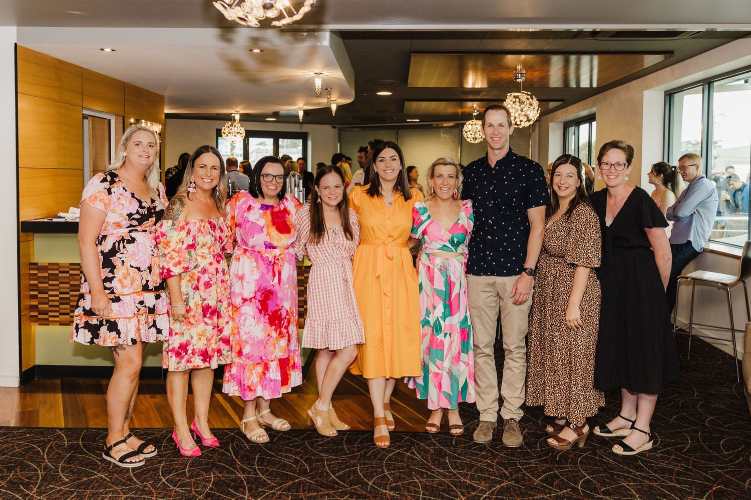 A group poses together at an indoor event wearing colorful floral dresses and summer attire.