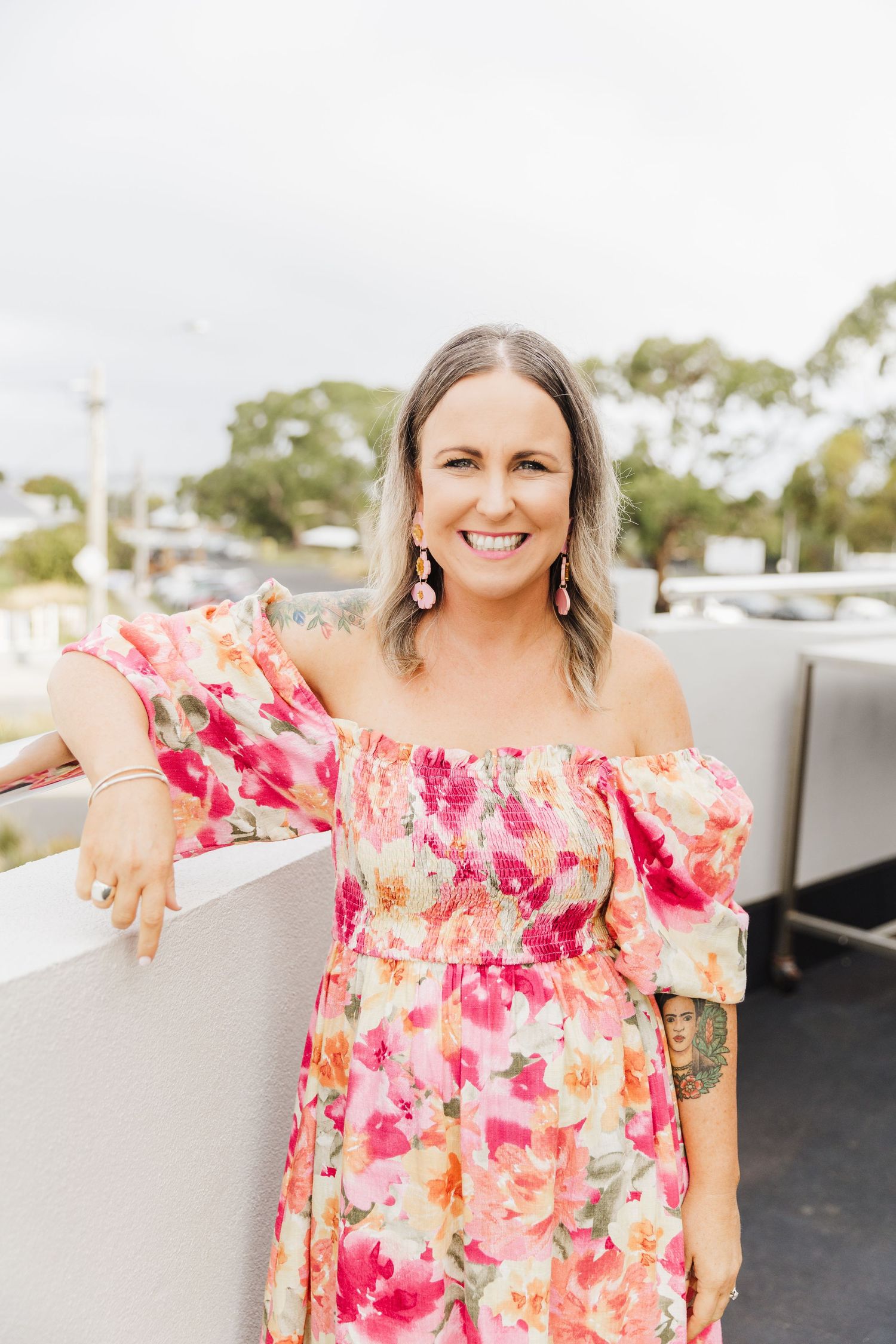 A person in a pink floral off-shoulder dress poses on an outdoor patio with a bright smile.