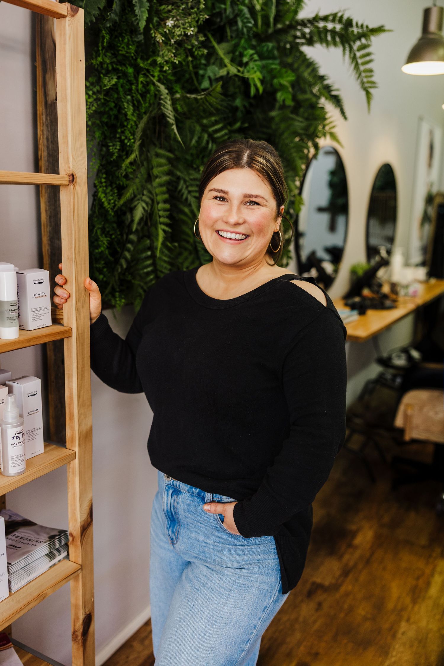 A person in a black sweater and jeans stands by wooden shelving displaying products in a bright retail space.