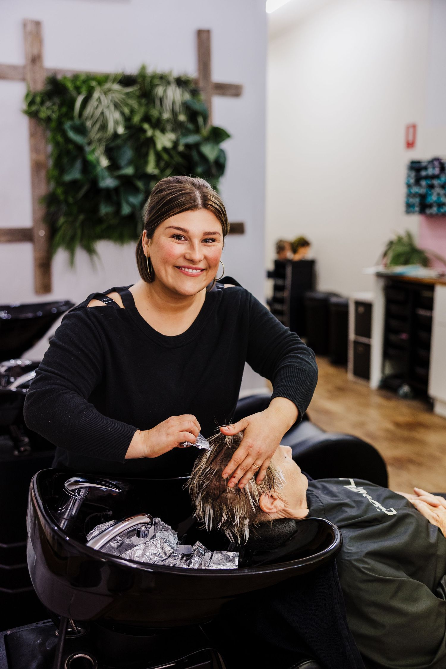 Hair stylist wearing black top washes client's hair in salon basin during professional hair care treatment.
