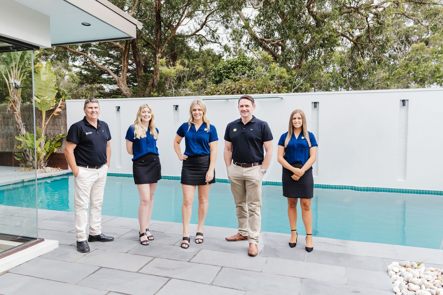 Five team members in matching navy and white uniforms stand in a line by a modern pool.
