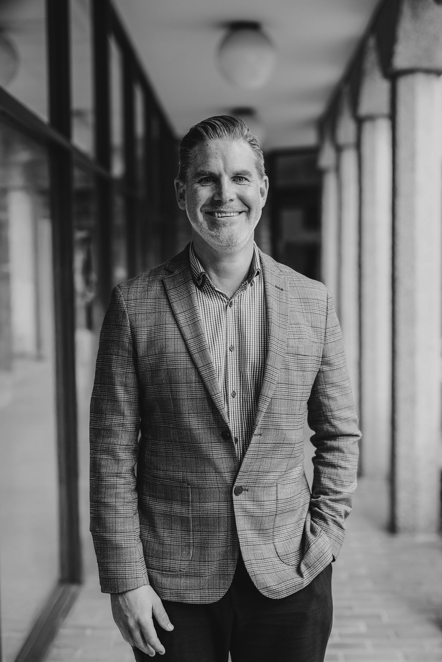 Professional black and white portrait of a person in a plaid blazer standing in an elegant columned hallway.