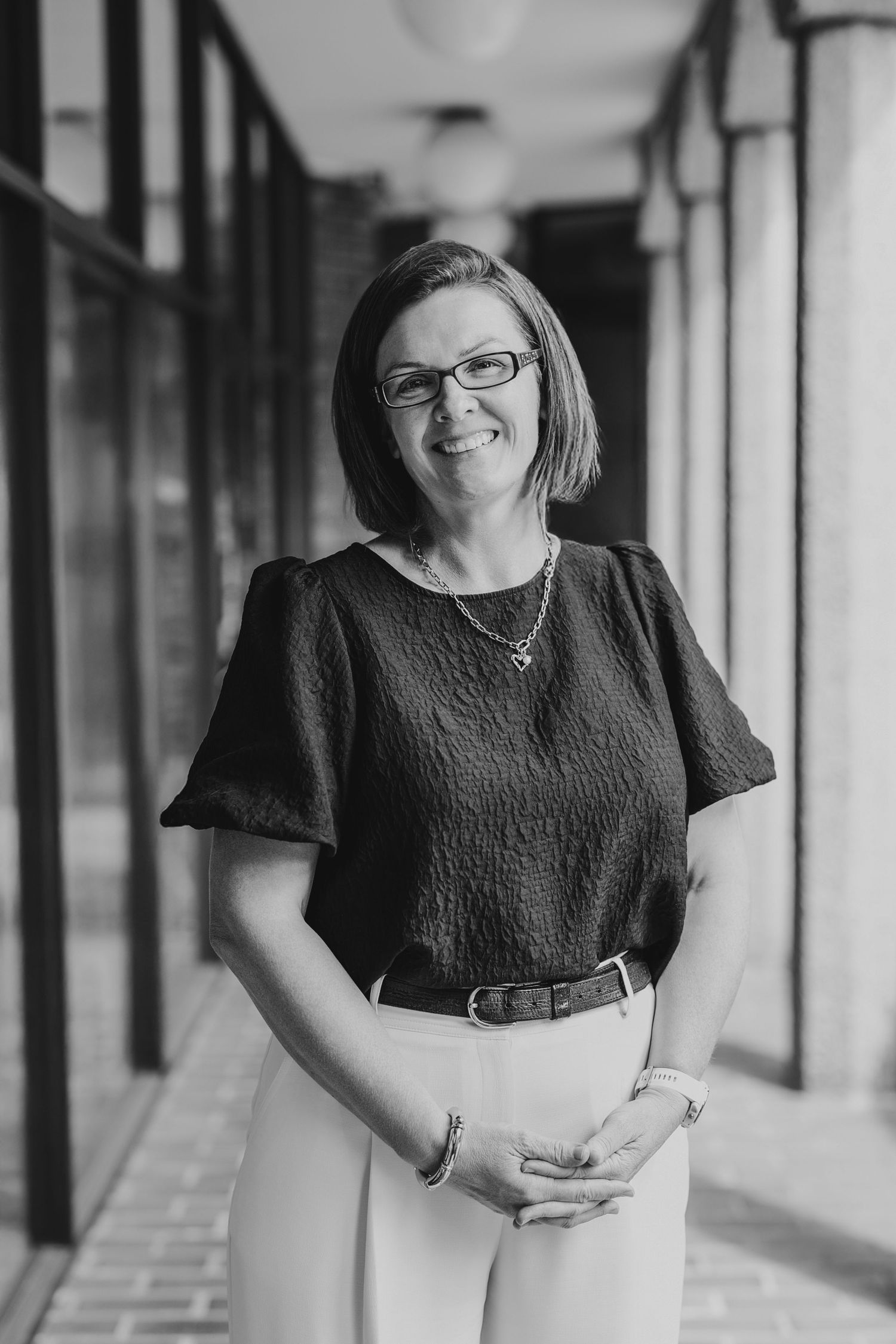 Black and white professional portrait of someone in glasses and dark top standing in a covered walkway.
