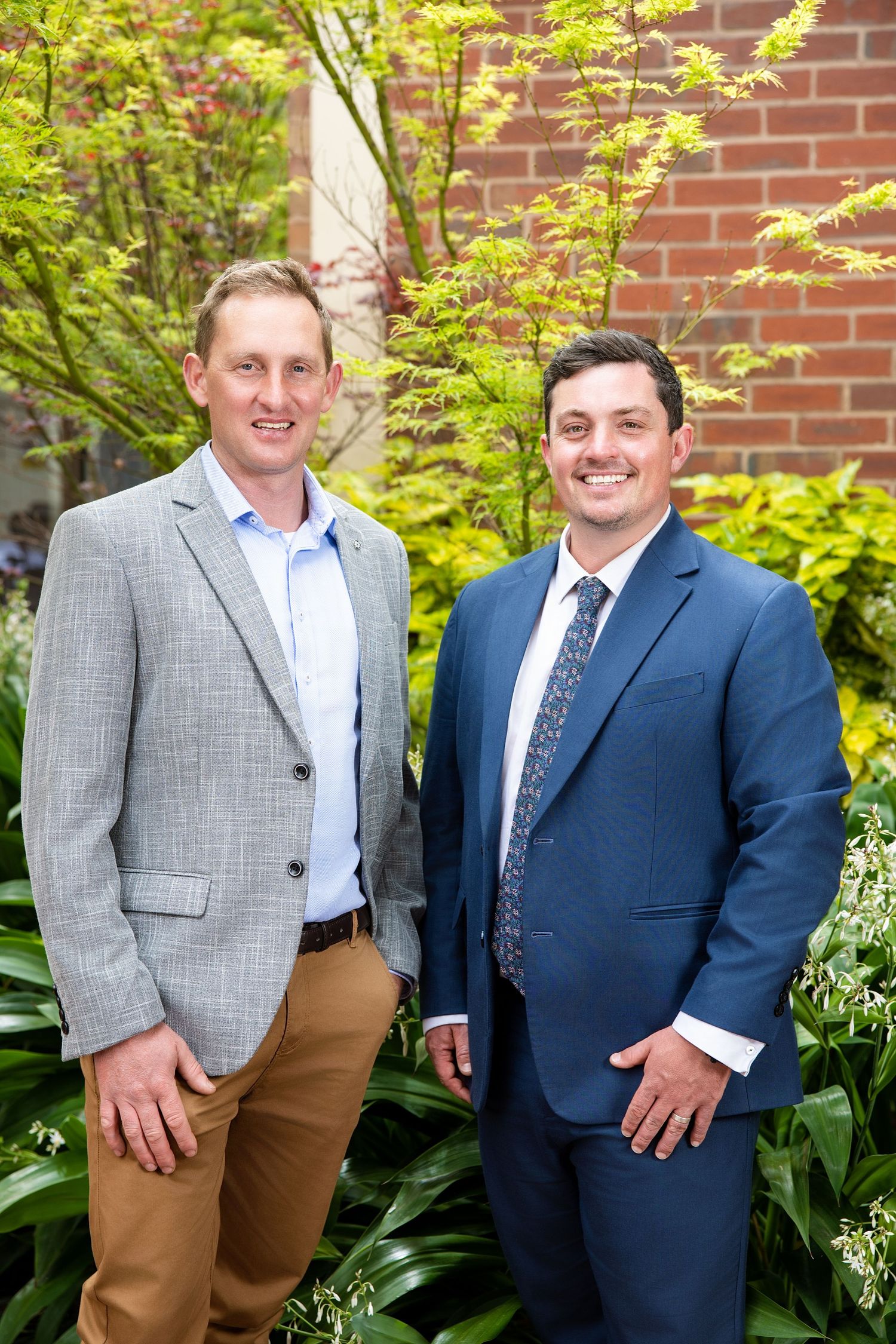 Two people in formal attire stand together against a brick wall with green foliage in the background.
