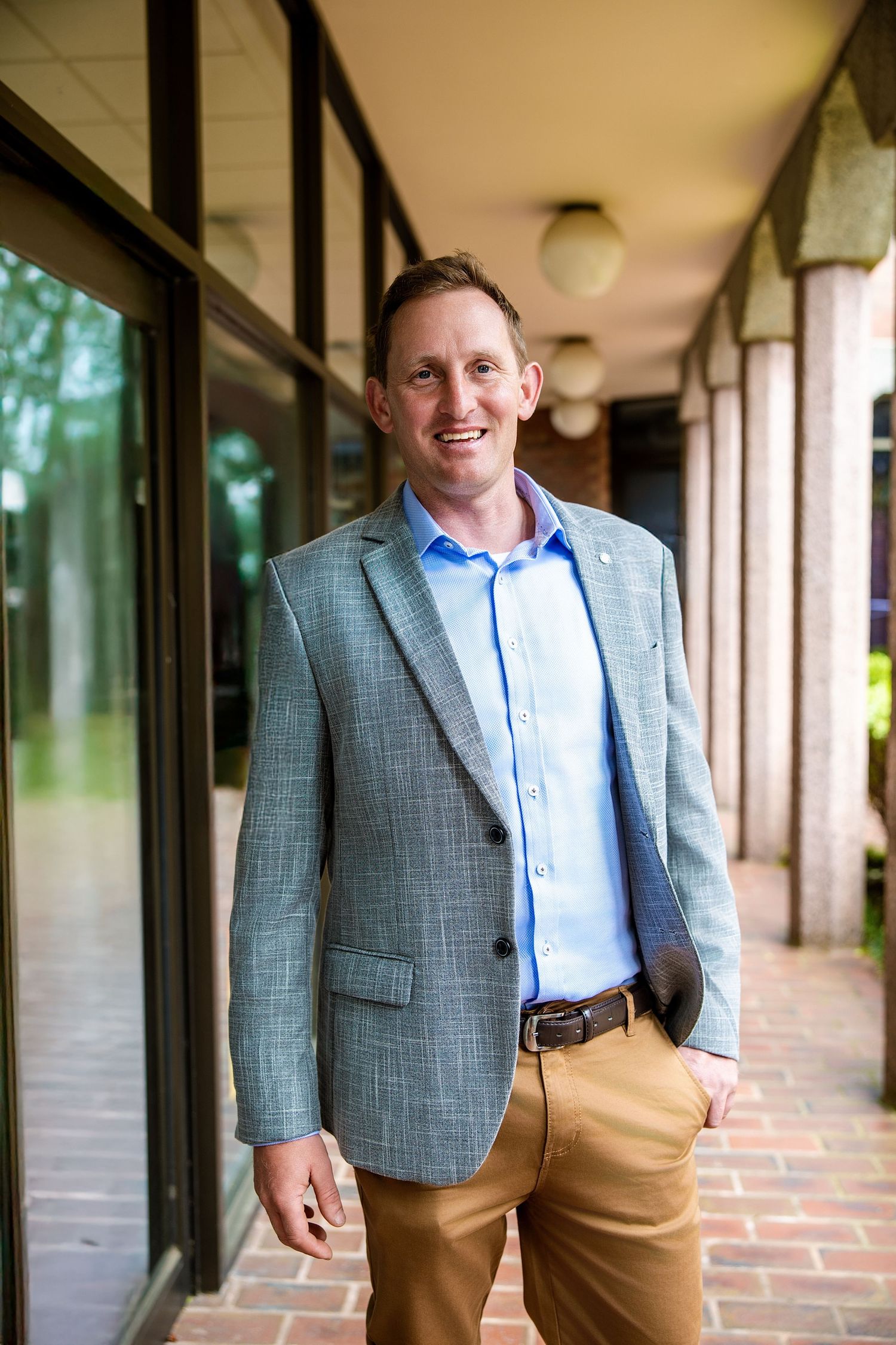 Professional wearing a gray blazer and khaki pants stands in an outdoor covered walkway with brick columns.