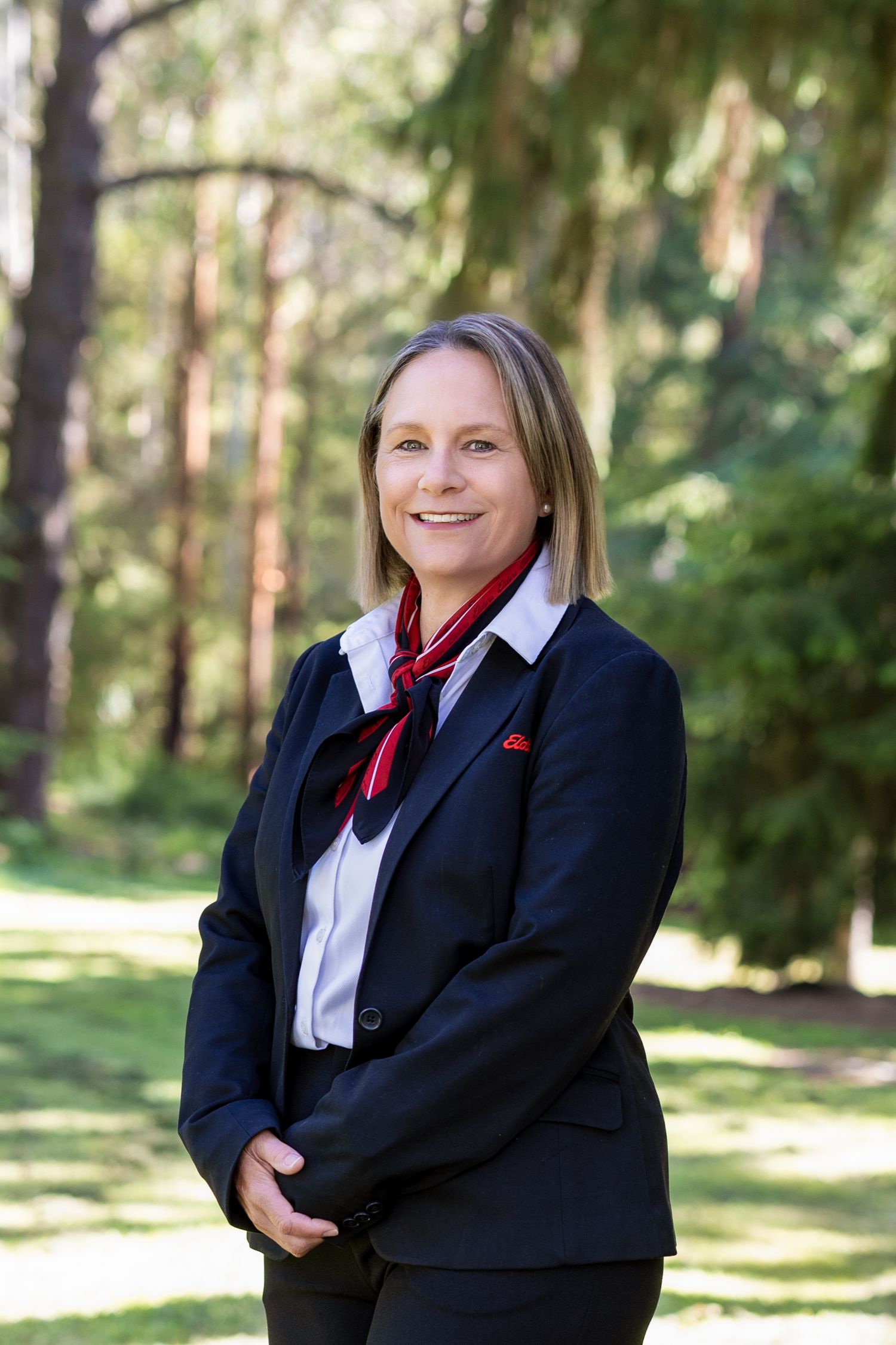 Professional wearing a black blazer and red bowtie poses in a bright outdoor forest setting.