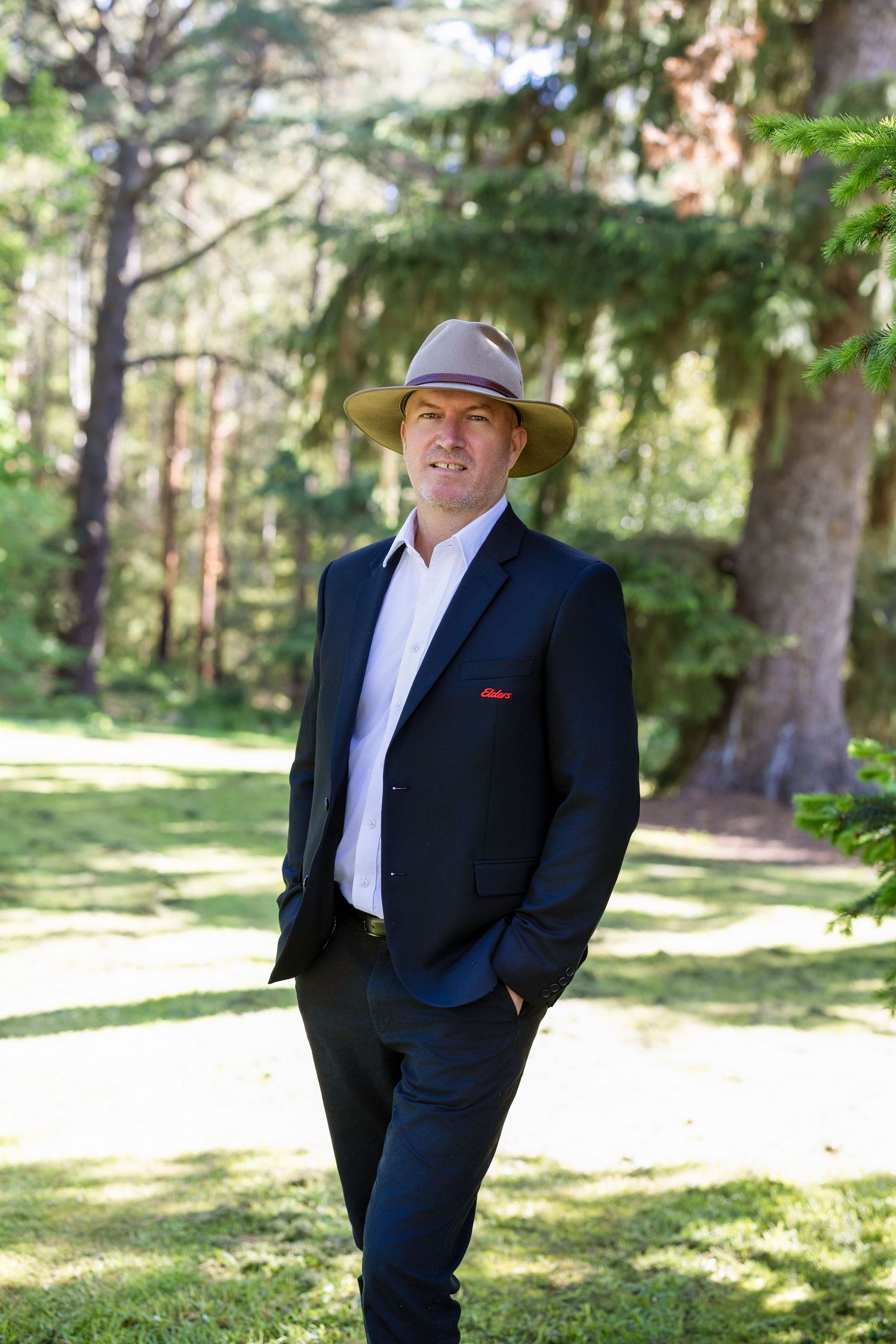 A person in a dark suit poses outdoors in a sunlit wooded park setting with tall trees in the background.