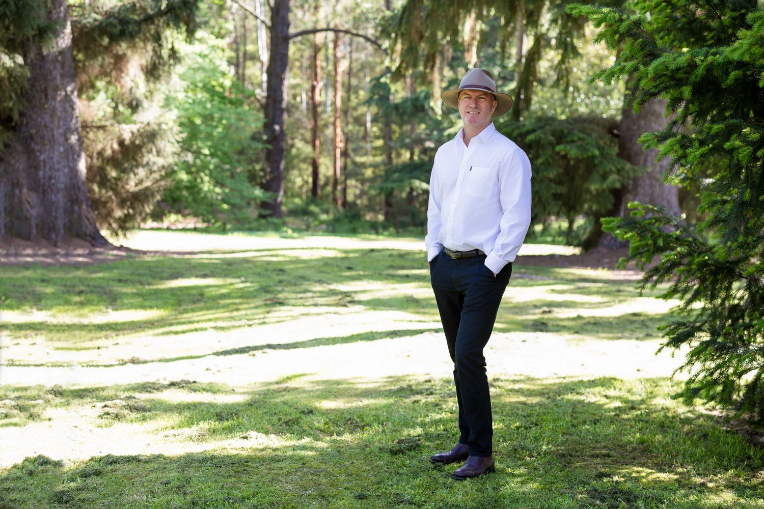 A person in white dress shirt and black pants stands in a sunny park surrounded by lush green trees and grass. Business Headshots Gippsland.