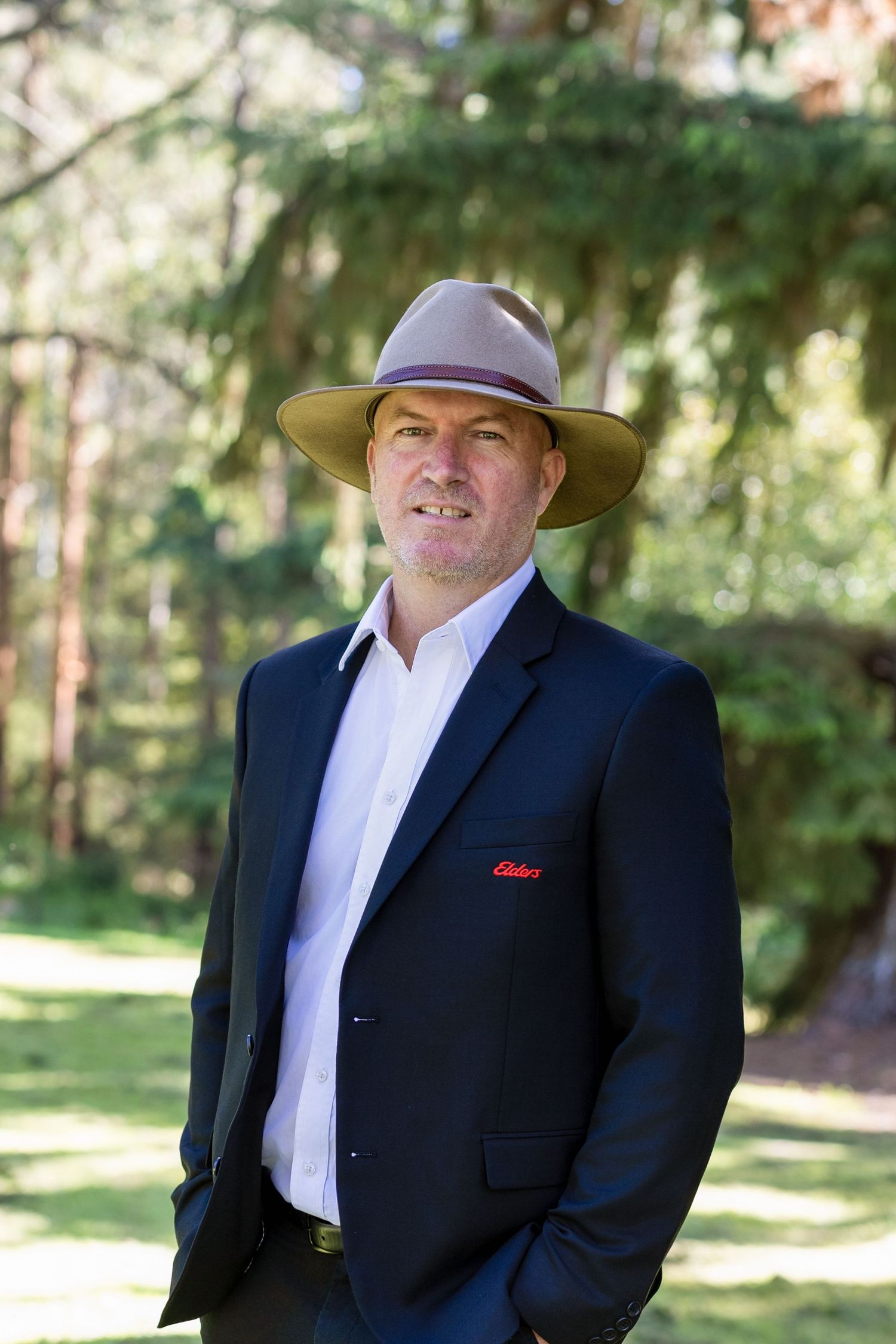 A person in a black suit and cowboy hat poses outdoors against a natural forest backdrop. Business Headshots Leongatha