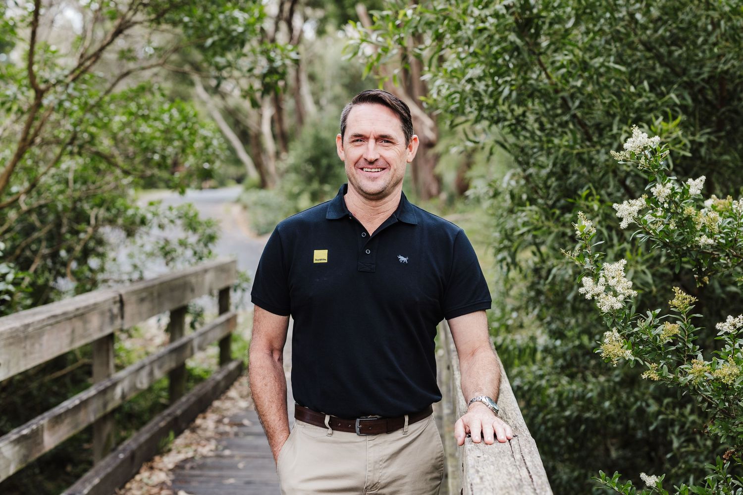 A person in a black polo shirt and khaki pants poses on a wooden boardwalk among flowering bushes.