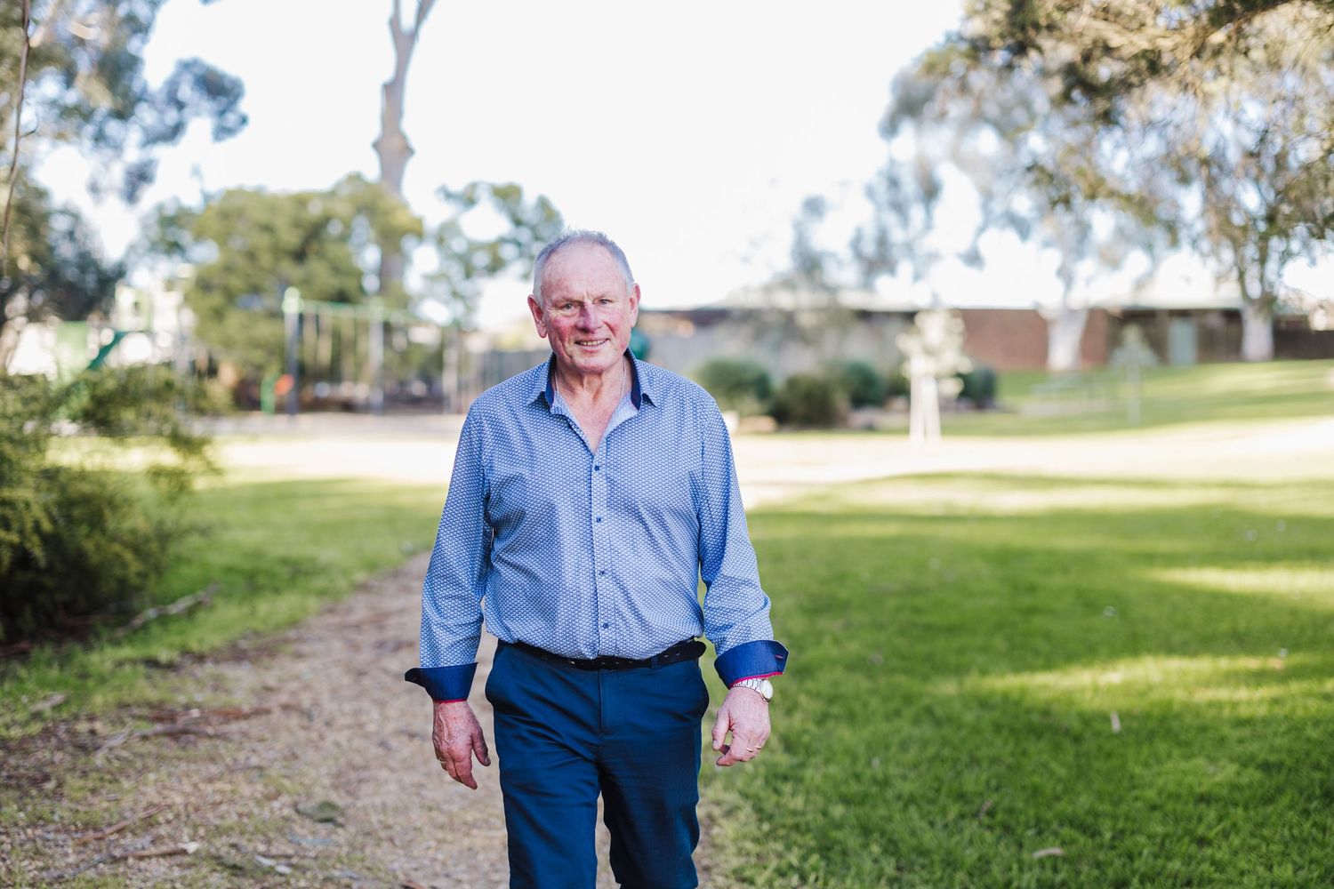 A person walks along a grassy path wearing a blue button-down shirt and navy pants on a sunny day.