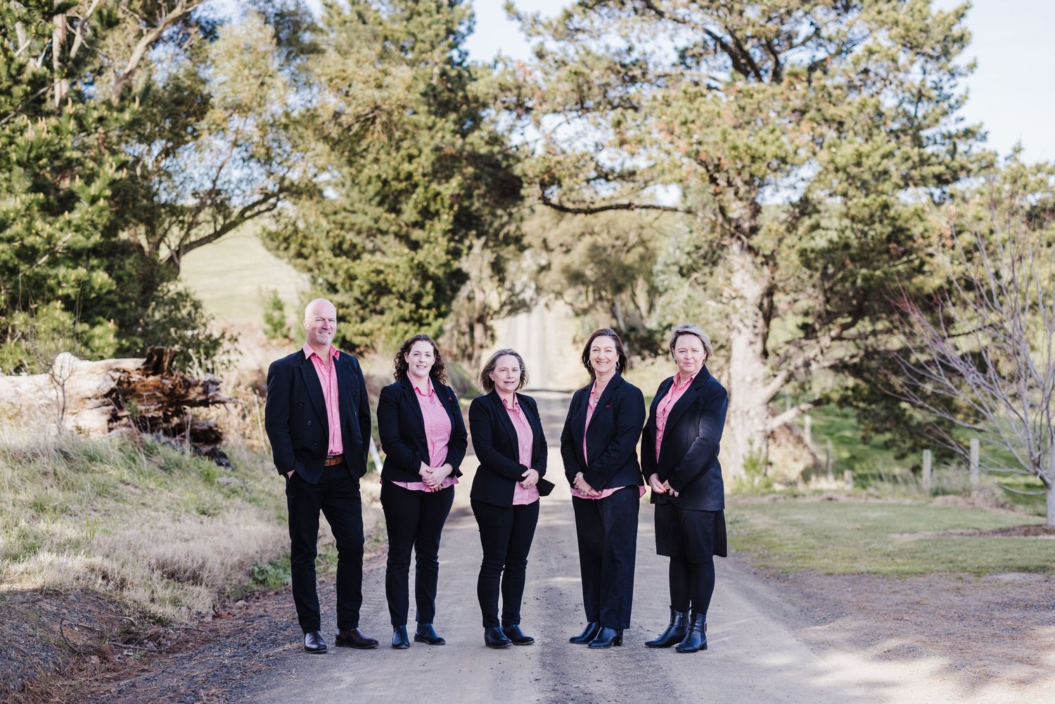 Group of professionals in business attire standing outdoors on a path surrounded by trees.