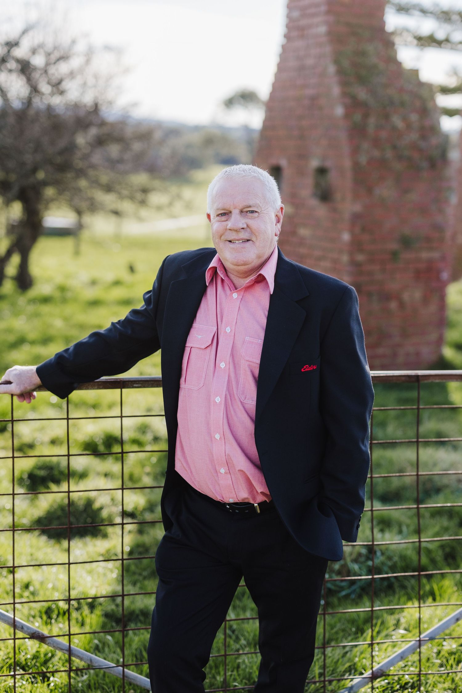Person in a suit and pink shirt standing by a rustic brick structure in countryside setting.