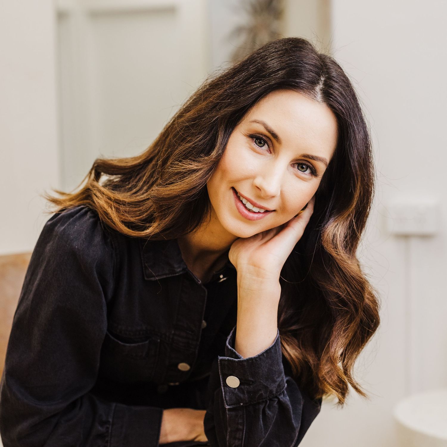 Person with wavy brown ombre hair smiles warmly while wearing a black denim jacket in a natural light setting.