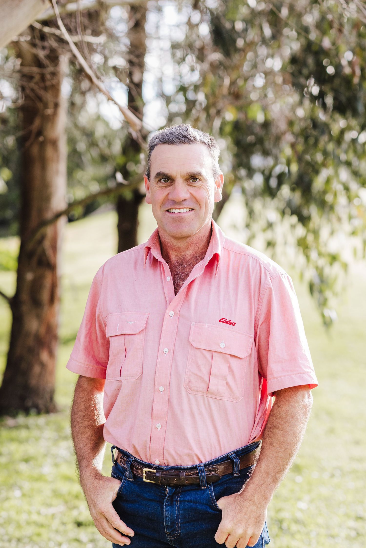 A person poses outdoors wearing a pink button-up shirt and jeans against a backdrop of eucalyptus trees.