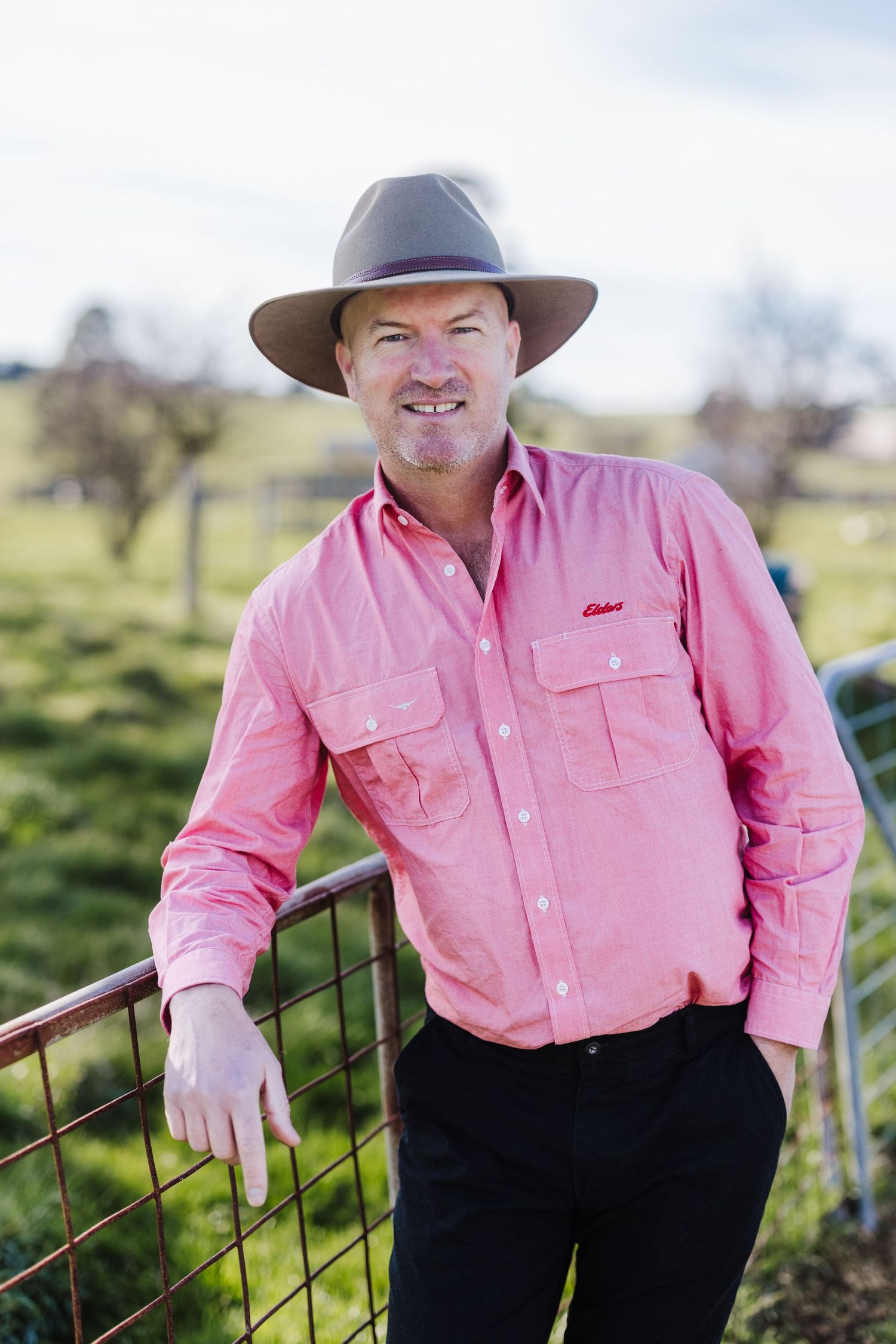 Person in a pink button-up shirt and brown hat leaning on a metal fence in a rural setting.