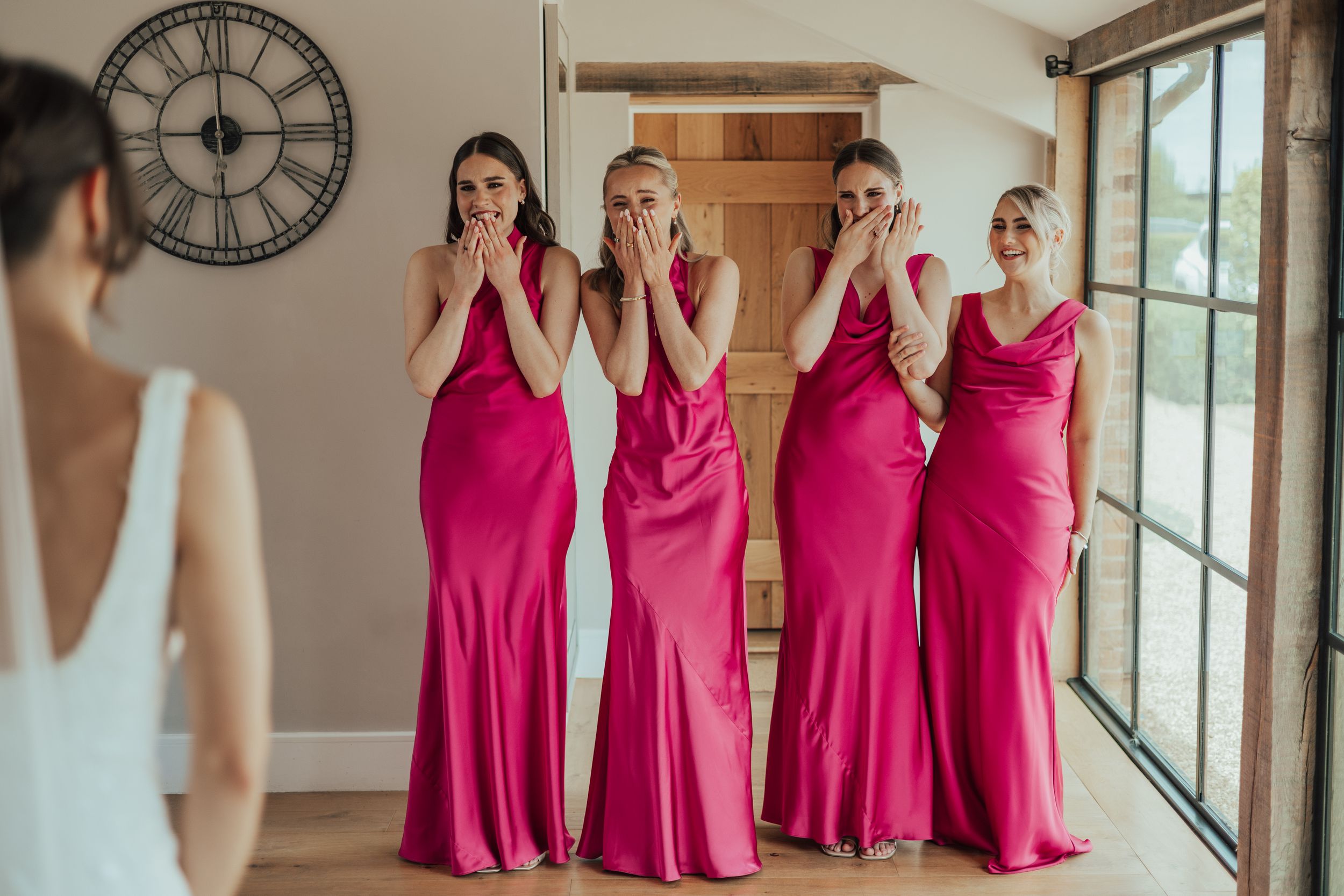 Four bridesmaids in pink satin gowns react emotionally seeing the bride in her white wedding dress.