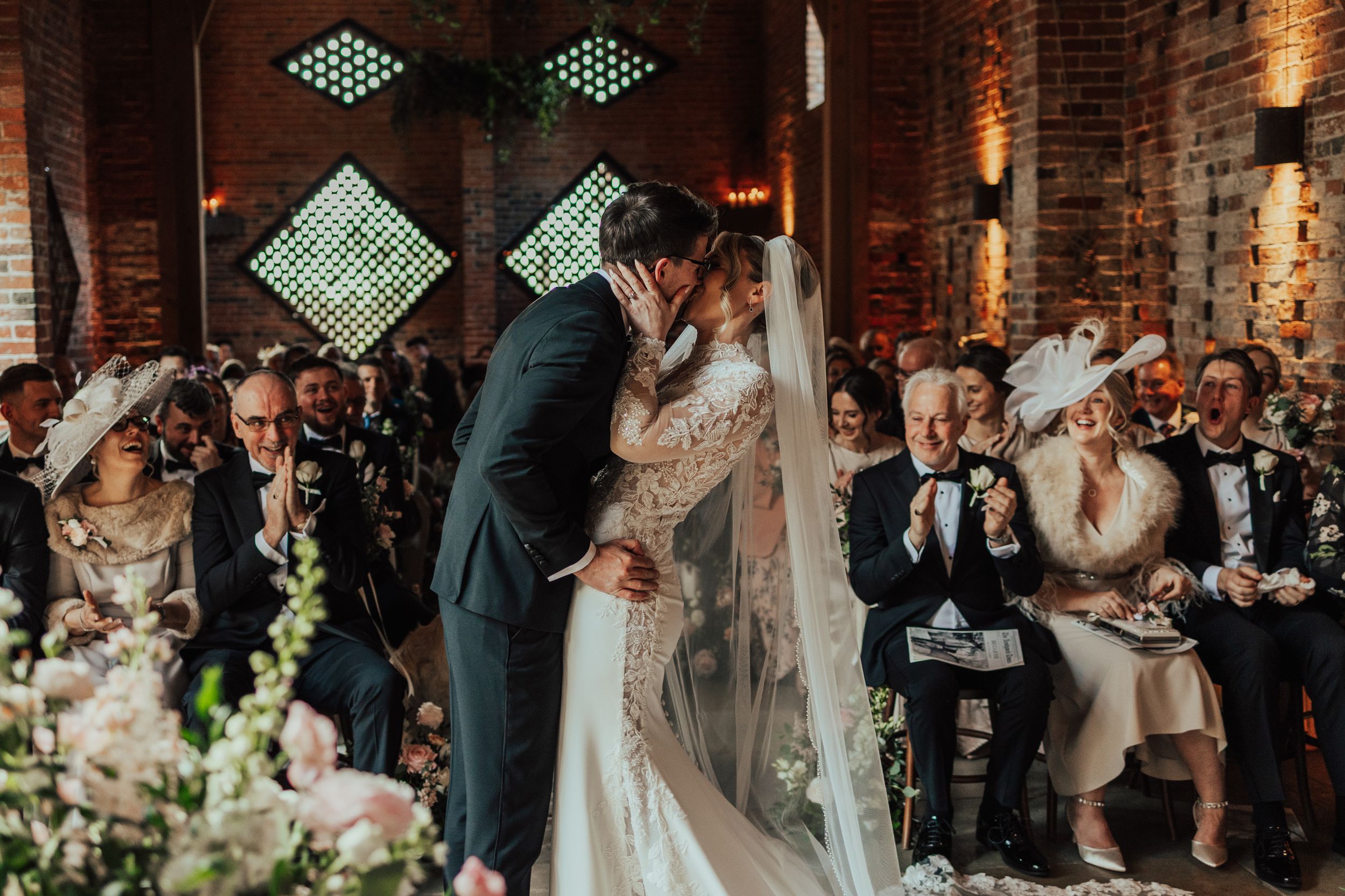 Bride and groom share first kiss at elegant barn wedding ceremony with guests applauding in background.