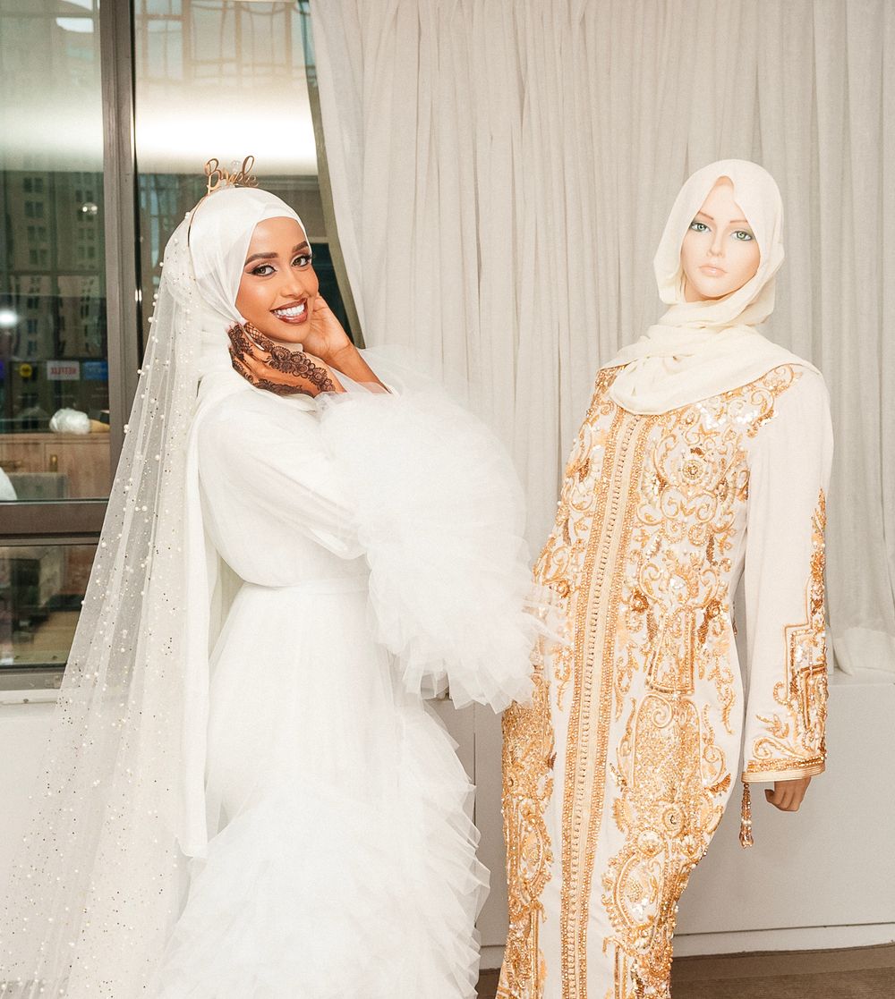Two women pose together in elegant hijabs and gowns, one in white bridal attire and one in a beige patterned dress.