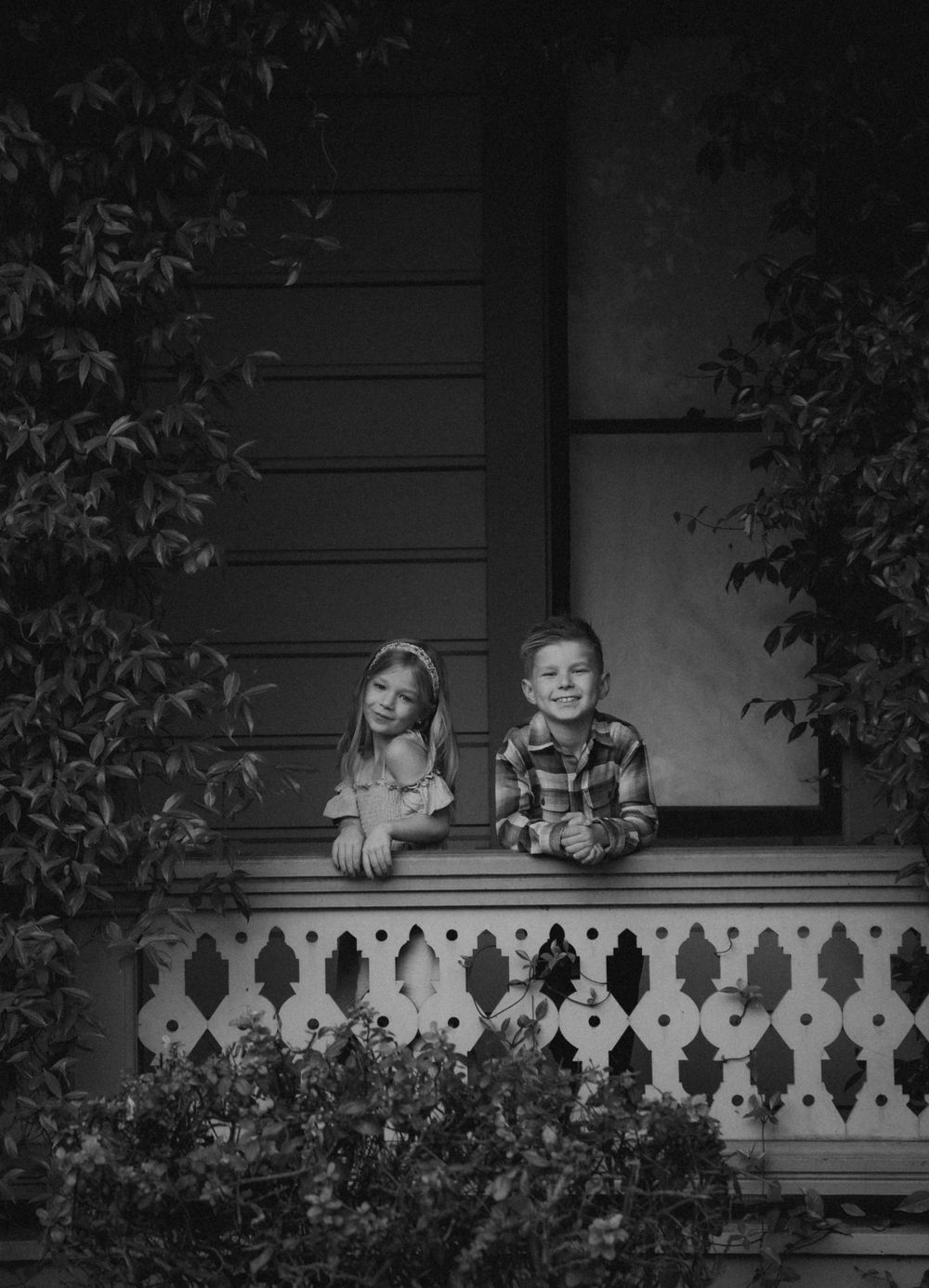 A black and white photo captures two children sitting on an ornate Victorian porch railing.