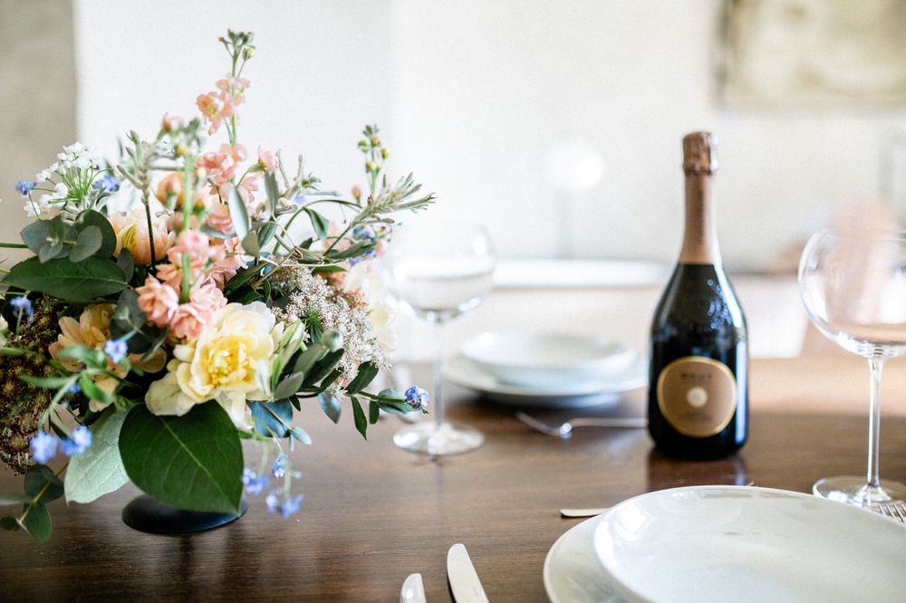 Elegant table setting with champagne bottle, wine glasses, and colorful floral centerpiece in peach, yellow and blue tones.