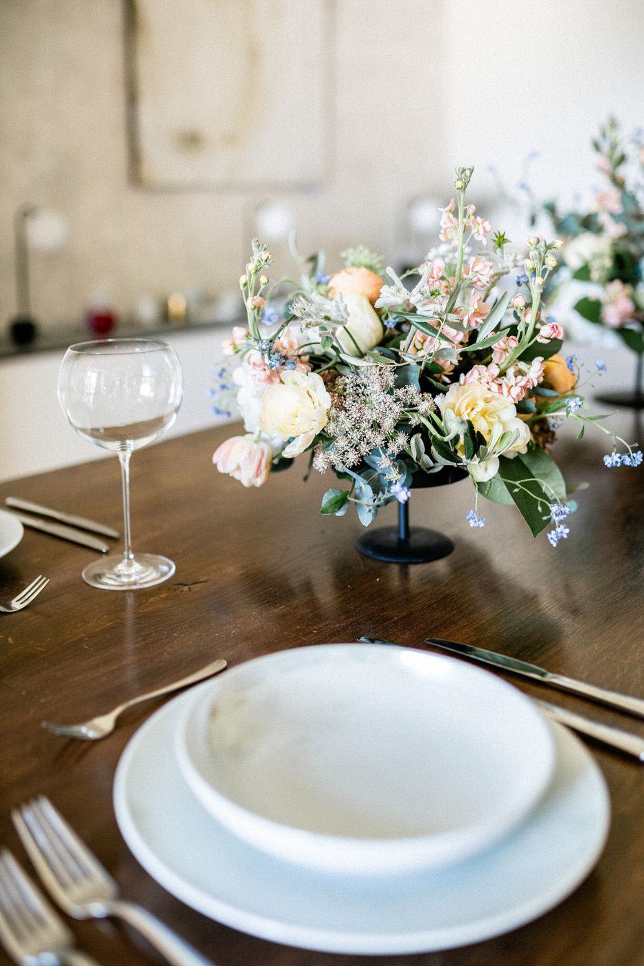 Elegant table setting with white plates, wine glass, and pastel floral centerpiece on dark wooden dining table.