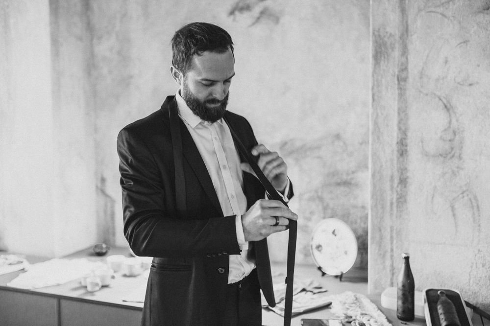 A man in a black suit and white shirt opens a wine bottle in a dimly lit room.