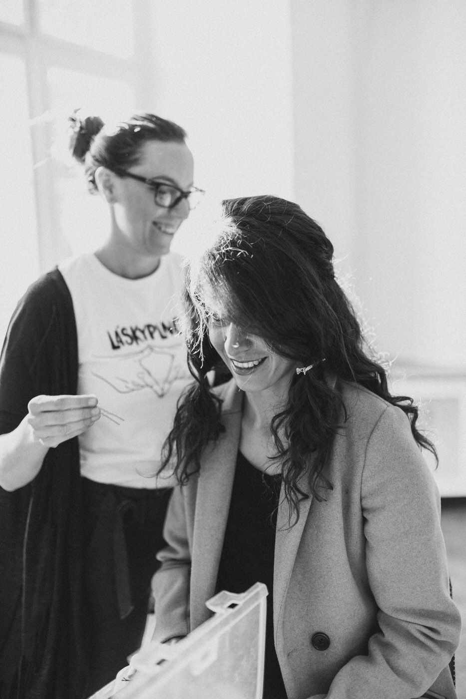 Black and white candid photo of two friends sharing a lighthearted moment together in a bright interior space.