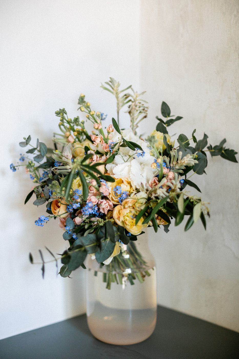Fresh garden style floral arrangement with yellow ranunculus, blue forget-me-nots and eucalyptus in frosted glass vase.