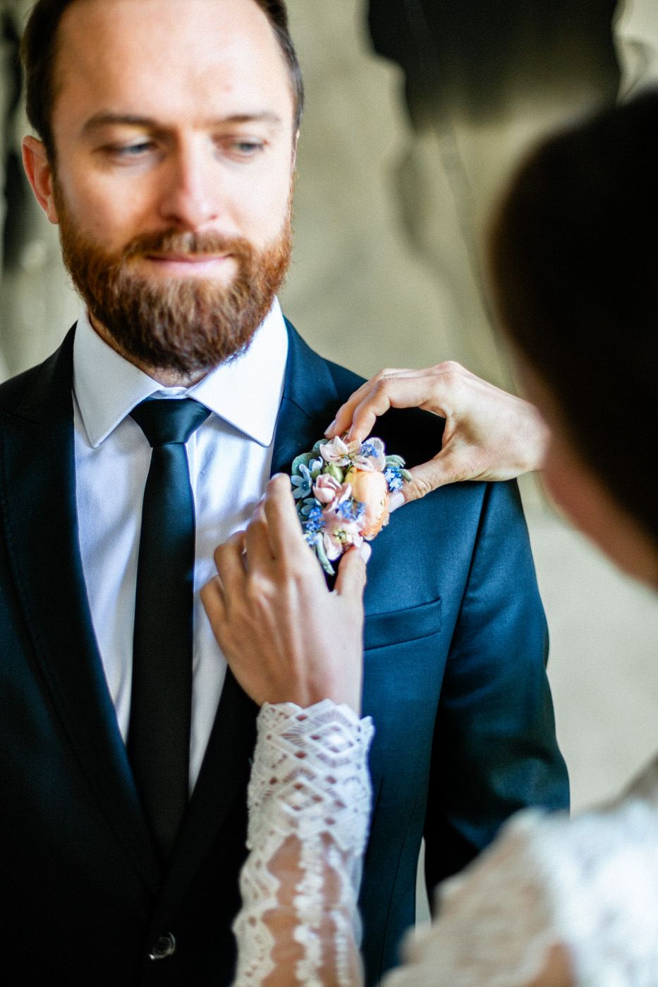 Delicate lace-covered hands pin a soft blue and pink floral boutonniere onto a navy suit jacket.