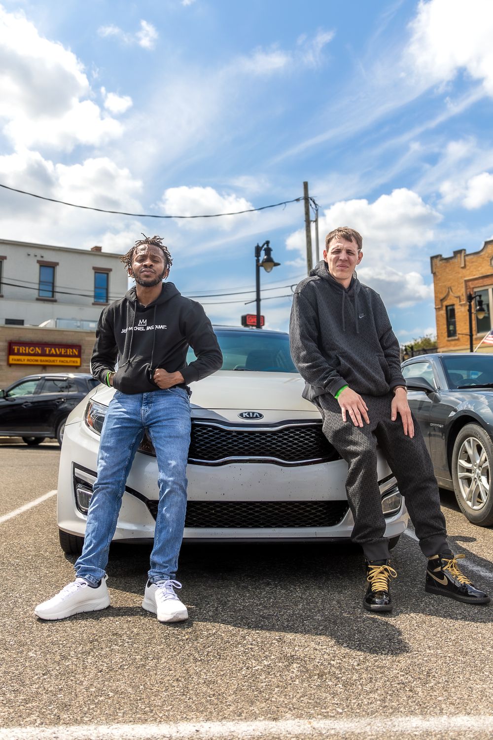 Two friends pose confidently in front of a white Kia car in a small town parking lot.
