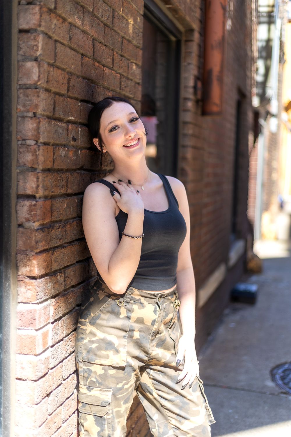 Casual portrait against brick wall wearing tank top and camo pants in urban setting.