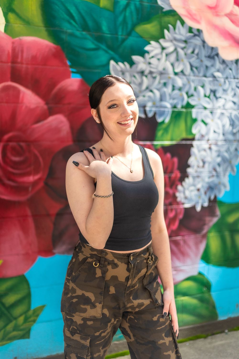 A vibrant floral mural serves as backdrop for a casual fashion pose in camo pants and black top.