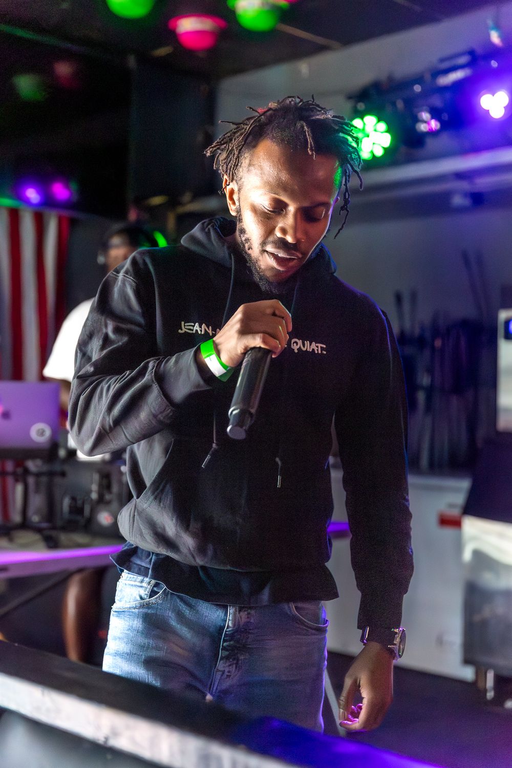 A performer in a black hoodie holds a microphone under colorful stage lights.