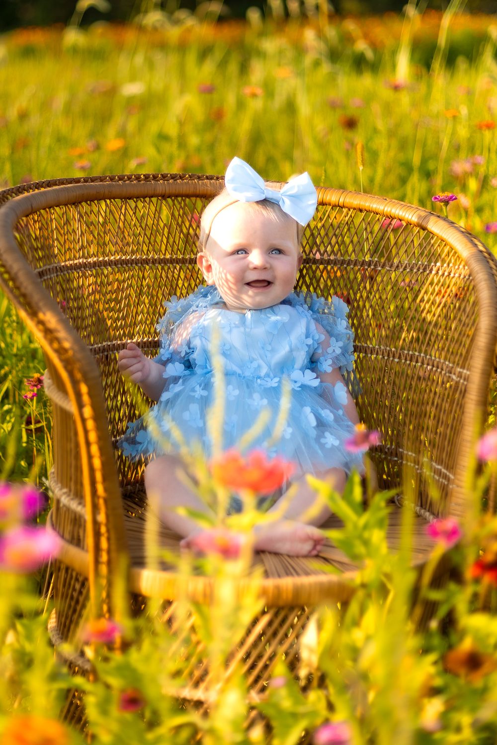 Smiling baby in blue outfit sits in wicker chair surrounded by wildflowers.
