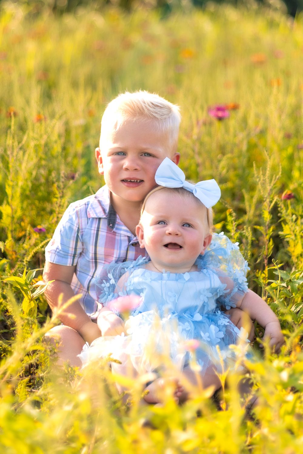 Two children sit together in a sunny wildflower field wearing coordinating blue and plaid outfits.